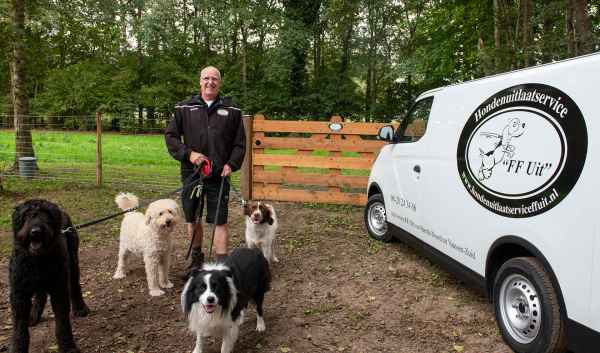 Bart staat te glimlachen terwijl hij de lijnen vasthoudt van vier honden van verschillende rassen en groottes. Ze staan op een grasrijk gebied met bomen op de achtergrond. Aan de rechterkant staat een witte bestelwagen met een logo en tekst die een hondenuitlaatservice adverteren, inclusief een cartoonhond en het webadres van de service.